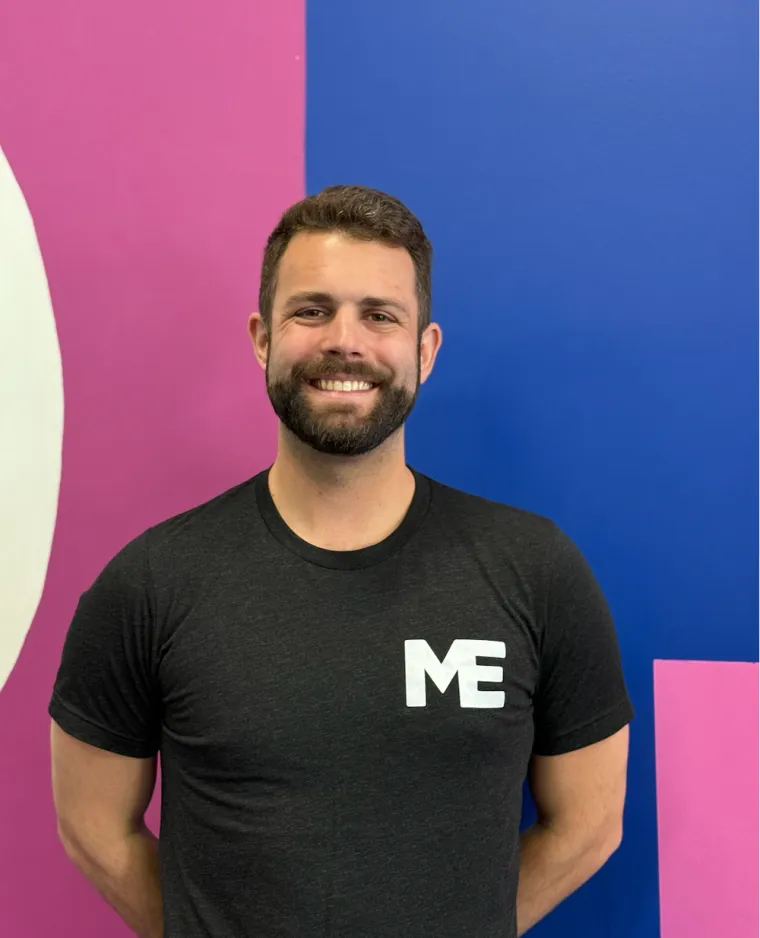 Elliot, strength and conditioning coach at Studio ME Fitness, smiling with arms crossed in a pink Studio ME shirt in front of the blue and white studio mural in Northeast Minneapolis.