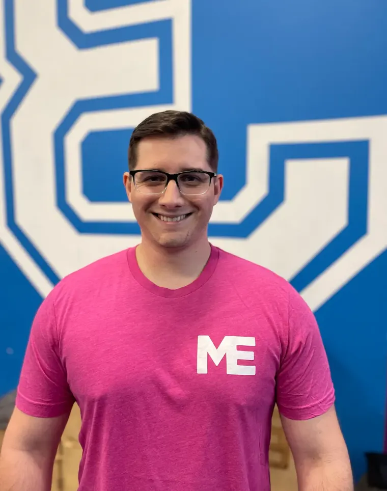 Luke, strength coach at Studio ME Fitness, smiling in a black Studio ME shirt in front of the pink and blue studio mural in Northeast Minneapolis.