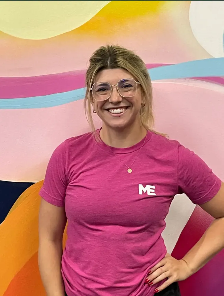 Shelby, coach at Studio ME Fitness, smiling with hand on hip in a pink Studio ME shirt in front of a colorful mural in Northeast Minneapolis.