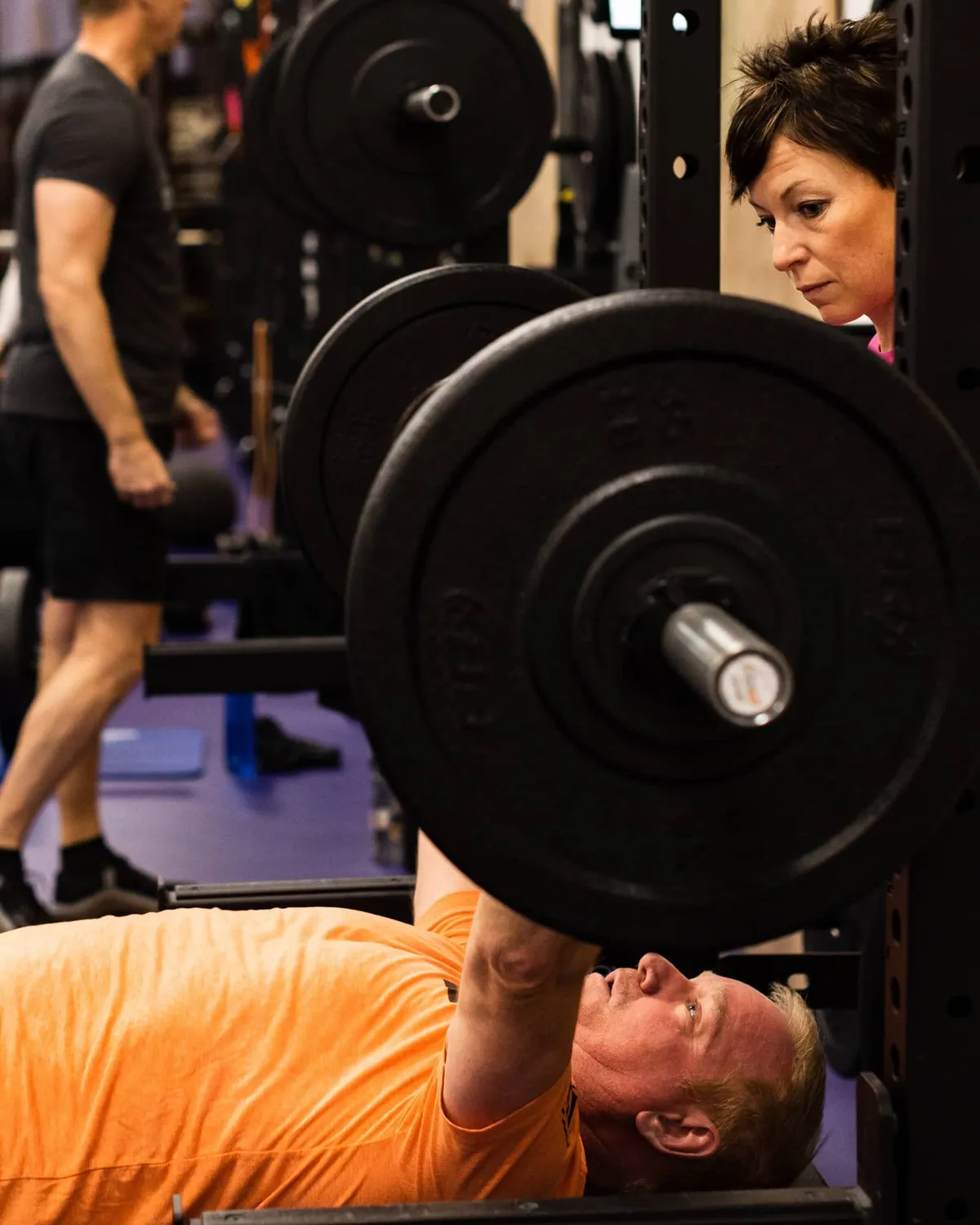 Studio ME Fitness coach in a pink shirt spotting a client during a barbell bench press at our Northeast Minneapolis strength studio, with another member training in the background.