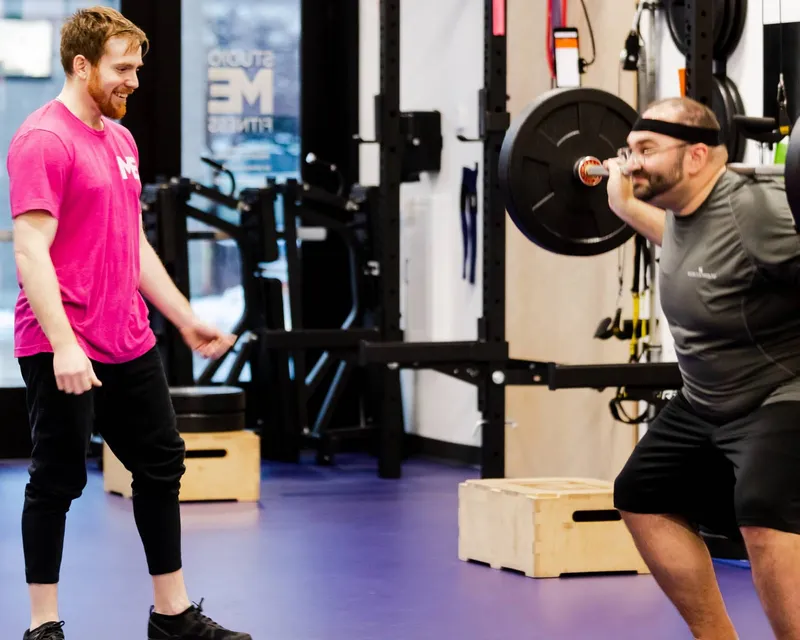 Studio ME Fitness coach in a pink shirt guiding a client through a barbell squat on the purple gym floor at our Northeast Minneapolis personal training studio.