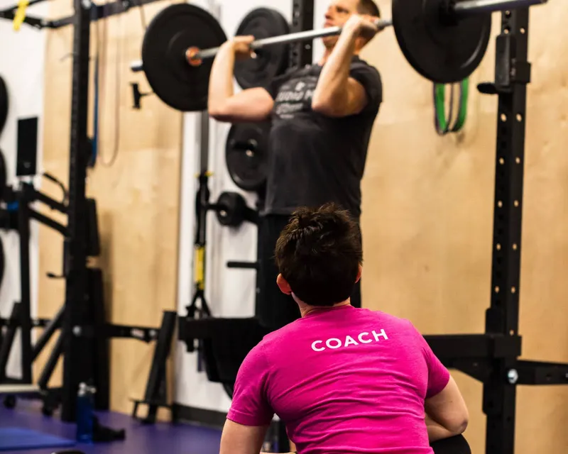 Studio ME Fitness coach in a pink COACH shirt observing a client performing a barbell back squat in the power rack during a semi-private training session in Northeast Minneapolis.