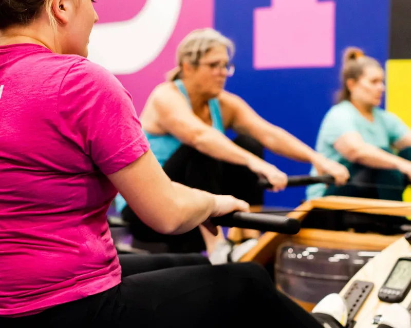 Three members rowing together during a small group training session at Studio ME Fitness in Northeast Minneapolis, with the studio's pink and purple branding on the wall behind them.