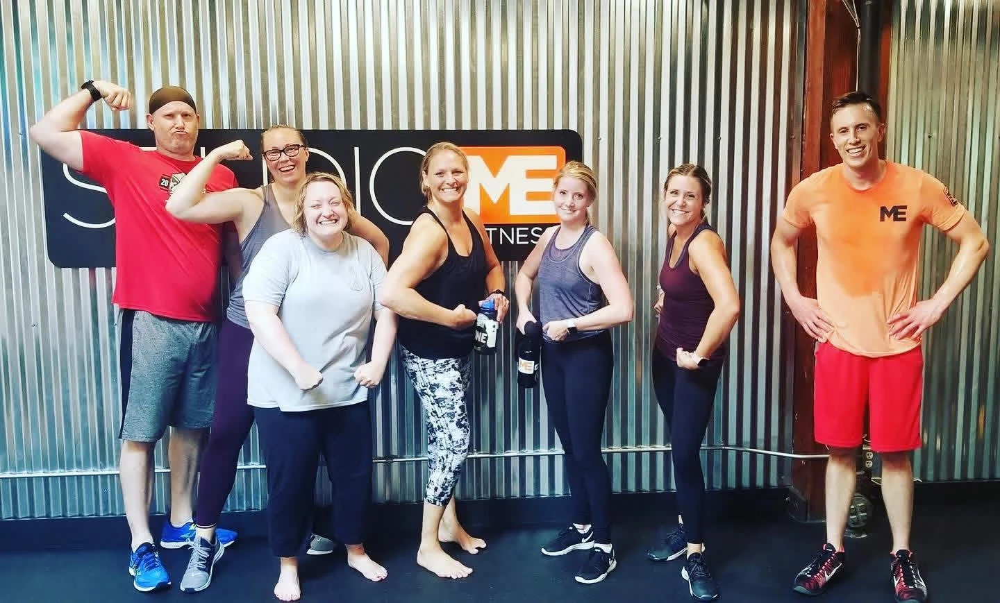 "Seven Studio ME Fitness members flexing and laughing in front of the studio's corrugated metal wall and orange 'ME' logo sign, in workout gear after a training session."