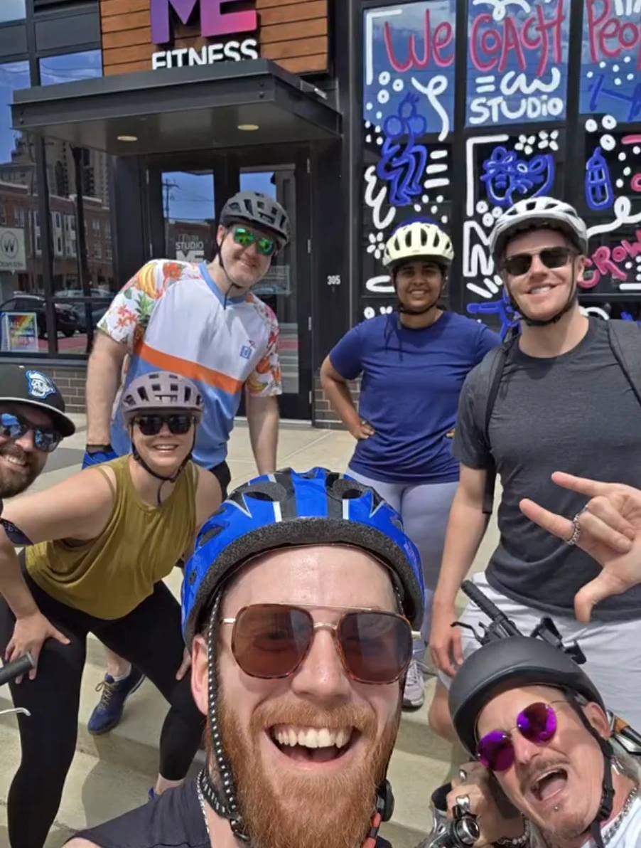"Ben (back left, in a floral cycling jersey and helmet) and Elizabeth (front, in an olive tank and helmet) smiling with a group of fellow cyclists outside Studio ME Fitness, in front of a colorful mural reading 'We Coach People.'"
