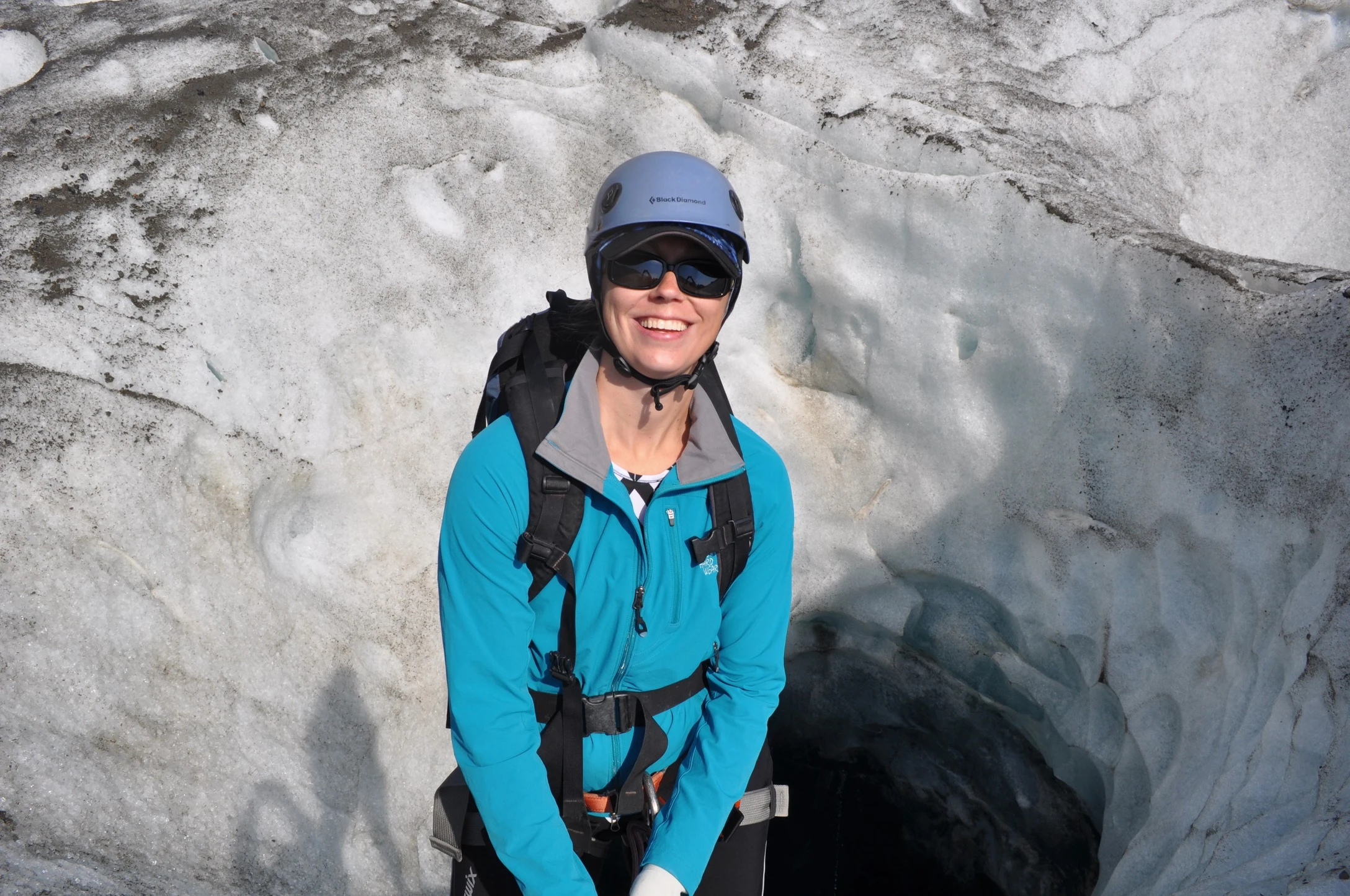 Elizabeth smiling in glacier gear and a helmet, standing in front of a glacial ice cave entrance carved into blue-white ice.
