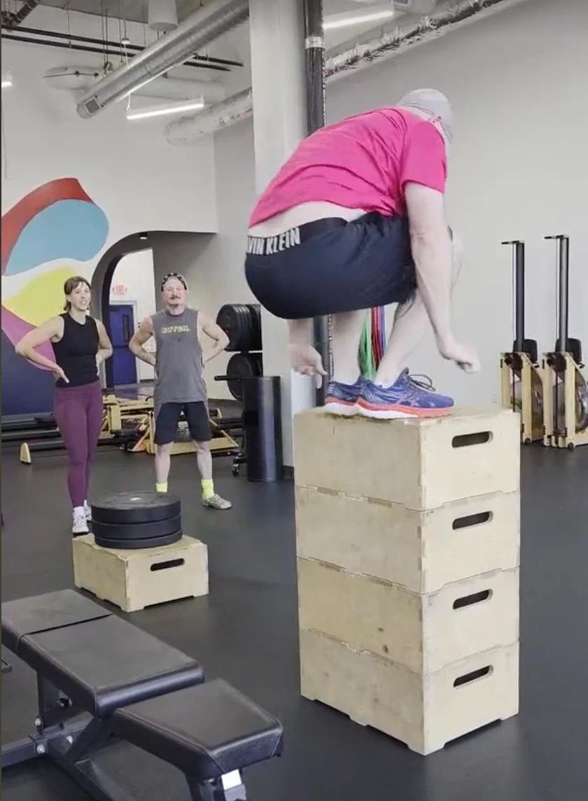 in a pink shirt mid-jump onto a stacked plyo box in the Studio ME gym, with a coach in a gray tank and a woman in maroon leggings watching nearby, rowers visible in the background."