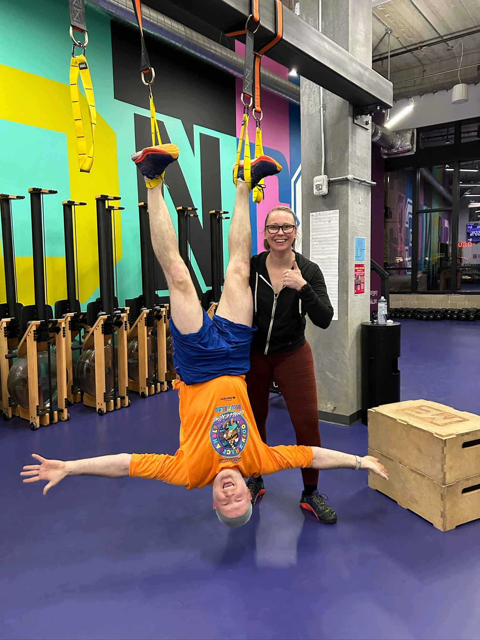 Ben hanging upside down from TRX straps with arms out and a big grin, while Elizabeth stands next to him giving a thumbs up, in the Studio ME gym with rowers and a colorful mural in the background.