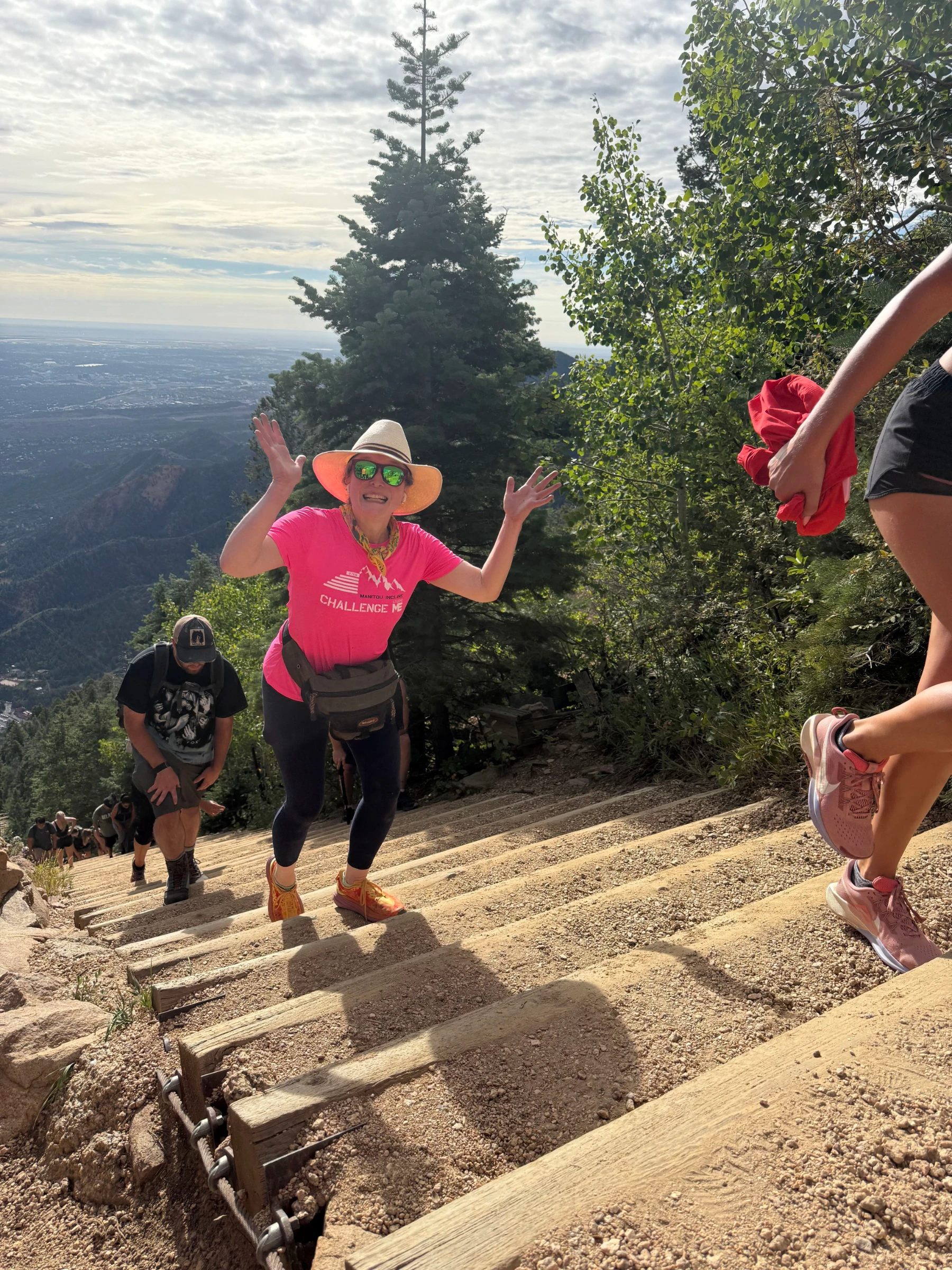 Melissa smiles and waves both hands mid-climb on the steep wooden steps of the Manitou Incline, wearing a pink Challenge ME shirt, with the Colorado landscape far below.