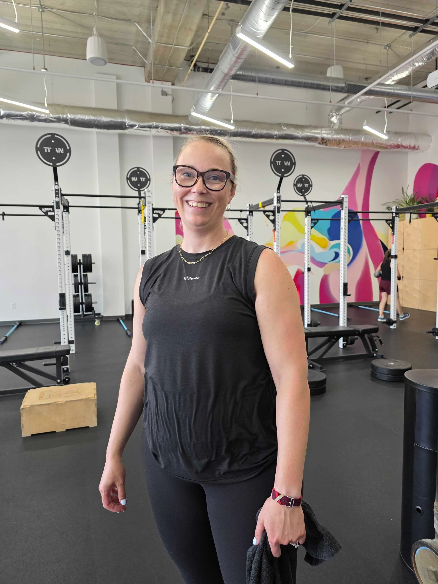 "Smiling woman in glasses and a black Lululemon tank standing in the Studio ME gym, with squat racks, weight plates, and a colorful mural in the background."